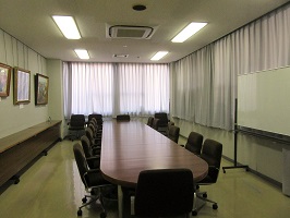 会議室の写真。中央に円卓、椅子17脚、ホワイトボード、予備の長机2台が映っている。